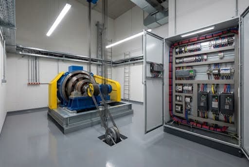 Casa de máquinas de elevador com máquina de tração azul e amarela sobre base metálica, cabos descendo ao poço e quadro de comando aberto com inversores e fiação